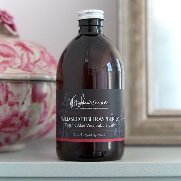Wild Scottish Raspberry Organic Aloe Vera Bubble Bath van The Highland Soap Company in amberkleurige fles, op een witte plank met roze bloemenvaas op de achtergrond
